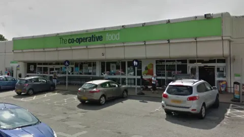 Google A large shopfront has green branding across the front which says The co-operative food. Several cars are parked in a car park in front of the shop.