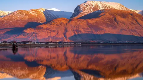 Ben Nevis, dusted with snow, rises above Caol, Inverlochy and Fort William. The peak and nearby mountains are reflected in the mirror-ike surface of Loch Linnhe.