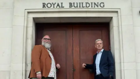 Plymouth City Council Councillor Chris Penberthy (left) and Councillor Mark Lowry (right) pictured in front of the main entrance to the Royal Building. The door is brown in colour and above there is a sign which reads the name of the building 'Royal Building'.