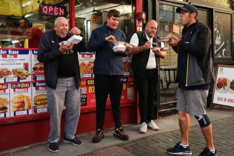 Getty Images Four men stood outside a takeaway shop eating out of polystyrene white containers. There are images of food behind it 