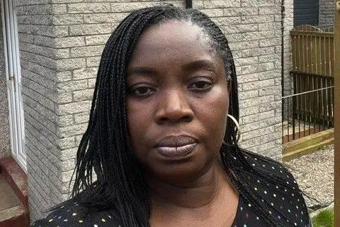 Jumoke Adebajo Jumoke Adebajo, a woman looking at the camera standing outside her home.