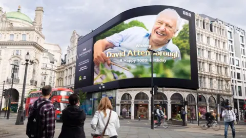 Billboard featuring a large image of Sir David Attenborough at Piccadilly Circus, with pedestrians and traffic in the foreground.