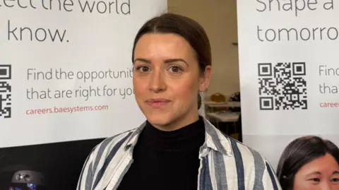 The photo shows a woman smiling at the camera. She has her hair tied in a brown bun behind her head. She's wearing a black top and a stripey shirt. There are banners with QR codes and text behind her. 
