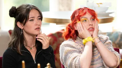 Studio Lambert/Euan Cherry/PA Two women look visibly shocked. The one on the left has dark hair, with fringe framing parts on her face and a small bun on one side. She wears a dark jumper with a silver zip detailing and a green necklace. The woman on the right has bright orange hair, has both hands to her face in a shocked expression and wears orange and red eyeshadow. She has a yellow bobble on her wrist and wears a striped knit jumper. The pair sit on red velvet chairs at a dining table.