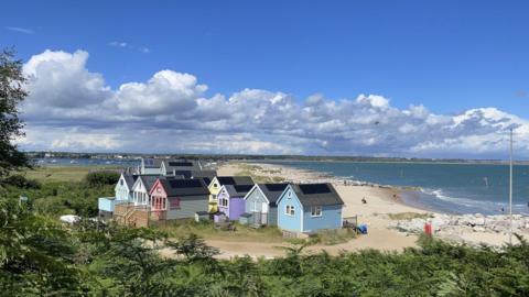 A group of twelve colourful beach huts are on Mudeford sandbank. On one side of the huts is the yellow sand on the beach and the sea, behind is Christchurch Harbour. In the distance you can see trees on the cliffs on the horizon. The sea looks blue on a sunny day and overheard are blue skies with white clouds.