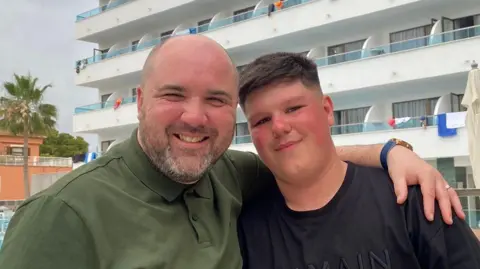 Charlie Hopkins There are two men in this photograph. On the left is Charlie Hopkins, the father of Morgan, who is smiling and wearing a green polo shirt. He's bald and has a beard. He has his arm around his son, Morgan, who is also smiling. He's wearing a black t-shirt and has brown hair.