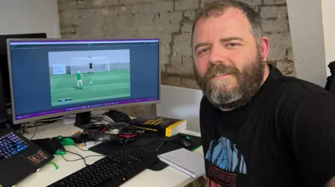 A man is sitting at an office workspace. His computer monitor shows a screenshot from a sports video game. He is wearing a black t-shirt and has a brown-grey beard with short brown hair