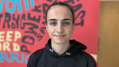 A young woman with dark brown hair, tied back, and wearing a black hoodie, stands and smiles at the camera. A large red display board is leaning against a door behind her.