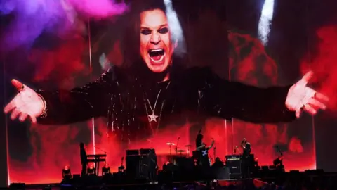 A band can be seen in red and black silouhette at the bottom of the image. On top the image of a man wearing black, chains around his neck, arms outstretched, dark eye make up and with a big smile 