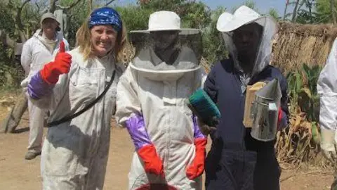 Rachel Monger Three people stand wearing beekeeping suits. One woman is not wearing the headwear and is doing a thumbs up and smiling, with a blue bandana on her head.