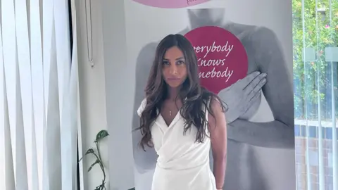 Jothie Walford pictured standing in front of a pink and grey sign that says Worcestershire breast unit haven. She is wearing a white dress and has long curly brown hair.