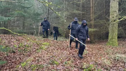 Four members of the search and rescue team in Hamsterley Forest. They are wearing black waterproof clothes and carry sticks. They are surrounded by dense trees. The ground is covered in brown leaves.