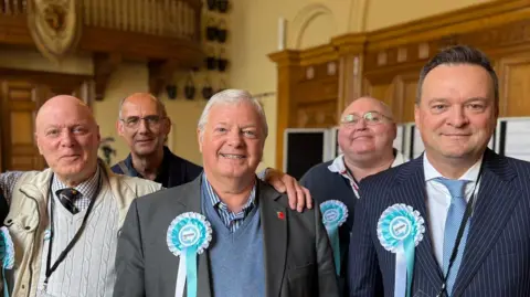 A group of men look into the camera. One on the right is wearing a blue pin-striped suit over a light shirt and blue tie. Some of the others wear blazers and jackets. Two at the back have glasses. Four have light blue and white rosettes with "Reform UK" written in the centre.