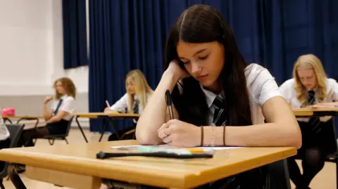 Getty Images A female secondary school student sits an exam in a large school hall concentrating on her paper. She has long brown hair and is wearing a white shirt and a school tie.