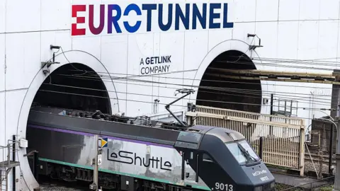 A LeShuttle train emerges from Eurotunnel - a white paneled structure with two dark tunnels. The train is dark blue and white and is attached to wires overhead. 