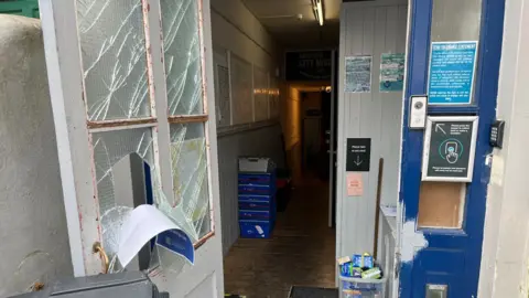 Lloyd Cummins Entrance door to the foodbank is open and shows four smashed windows, one of them with a hole. The hallway to the building is on display.