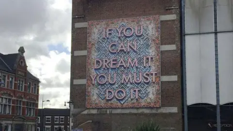 Robin Stott/Geograph If You Can Dream It You Must Do It artwork in Luton. The artwork is on a brick building, surrounded by other brick buildings. The working is in a red colour, with a background in blue and orange colours