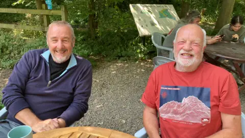 Both men in the seventies with grey hair and trimmed grey beards. Steven on the left is wearing a purple zipped collar jumper with light blue collar on the inside and David is wearing a red t-shirt