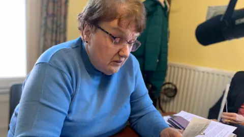 A woman, with short brown hair, is wearing a blue jumper and glasses while sitting on a chair reading an article from a newspaper