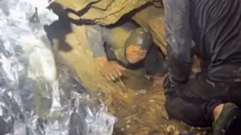 A man's head and arms emerge from a flooded cave with rescuers around him