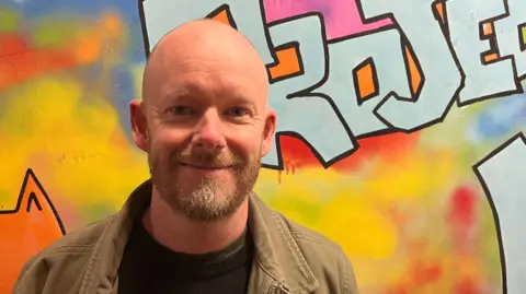 Ollie Conopo/BBC A man with a beard stands in front of a wall with graffiti art on it.