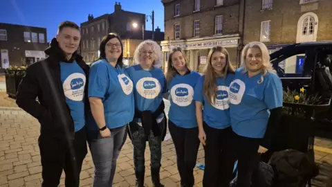 Sue Ryder The group of people with a man in the left and all the rest women, wearing blue Sue Ryder T-shirts, smiling for the camera.