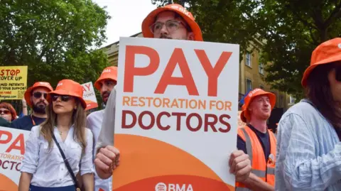 Resident doctors outside holding placards. They are wearing orange sun hats with a logo saying BMA. One holds a placard that says pay restoration for doctors. 