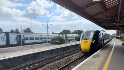 GWR A yellow and dark green GWR train alongside one of the platforms at Castle Cary station. Across the tracks in the distance, at another platform, is a grey SWR train. There are blue skies above with white clouds.