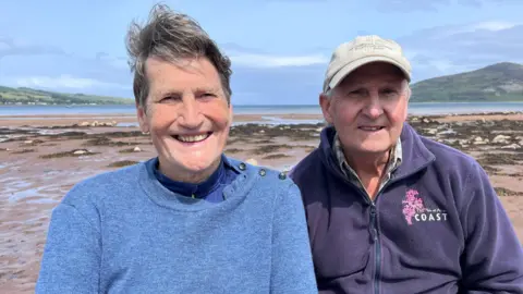 Don MacNeish in a blue jumper sits alongside Howard Wood, in a purple fleece and khaki baseball cat, on the beach at Lamlash bay, Arran, with the Holy Isle behind them