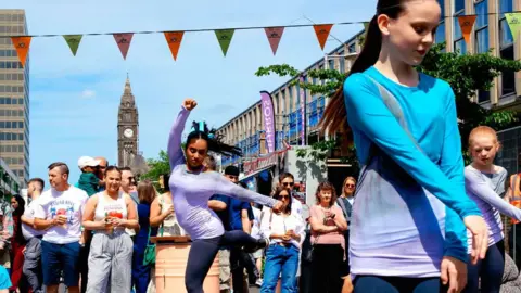 Stuart Whincup/BBC Crowds watch on a sunny day as a group of young dancers perform in the street. There is green and orange bunting drapped across the street and many people are wearing T-shirts. The tall, town clock can be seen against a blue sky background. 