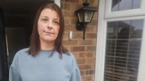 A woman stares into the camera from her doorstep. She has shoulder-length brown hair and is wearing a grey jumper.