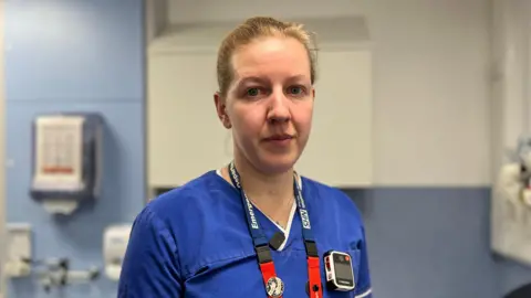 BBC Hannah Freer, a senior nurse at the Queen's Medical Centre in Nottingham. Hannah is a white woman in her later 30s, she has blonde hair tied back in a bun, she is wearing blue scrubs and has a body-worn camera clipped to her T-shirt.