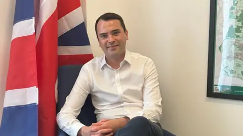 Amy Holmes/BBC A picture of the Conservative MP for Broxbourne Lewis Cocking, he is sat in a chair in his office, with his legs crossed. There is a Union flag next to him, and you can see part of a map of his constituency to his left. He is clean shaven, with short dark hair, and is wearing a white shirt with black trousers.