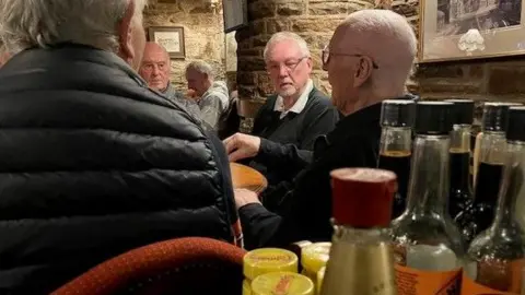 Um grupo de cavalheiros na casa dos oitenta está sentado conversando em uma mesa de pub. As tampas de algumas garrafas de condimentos estão em primeiro plano.