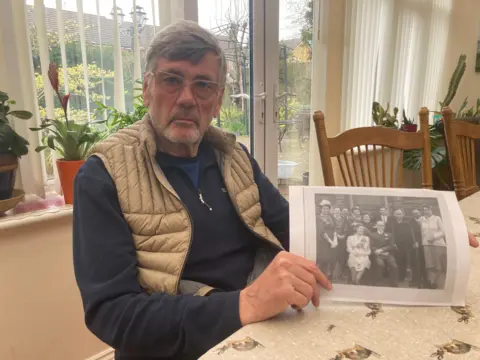 Bob Ainsworth sits at a table holding a black and white image from Len and Peggy Tasker's wedding day, which his own parents had attended. He is wearing glasses, a dark top and light brown gilet.