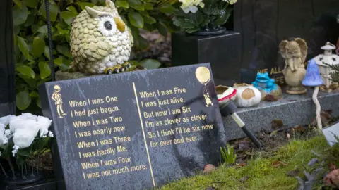 PA Media A dark marble memorial plaque with a poem and illustration of a child and Winnie The Pooh. There are toys and little statues scattered around. A statue of an Owl stands on top of the plaque, which sits among grass and trees.