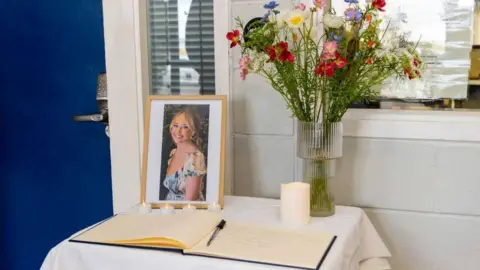 Dundalk Institute of Technology A memorial table featuring a framed photo, a glass vase with colourful wildflowers, a lit white candle, and an open book for messages, all arranged on a white cloth near a blue door and a window.