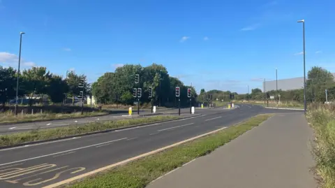 BBC A picture of the road with traffic lights and a yellow stop sign and grass verge. Next to this is a grey pavement. 