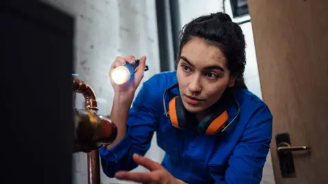 Getty Images Young woman in boiler suit with ear defenders around her neck looking with a torch at a boiler