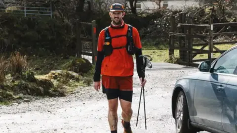 Sam Carden / Climb South West George Bell-Starr walking along a rural track, wearing an orange t-shirt and shorts, hiking boots, a hydration vest and black baseball cap. He's got a full dark beard and is carrying two Nordic poles in his left hand.