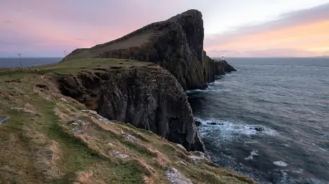 Matthew Boyle Steep sea cliffs extend toward a distant lighthouse at sunset, with waves crashing below and soft, pink light in the sky.