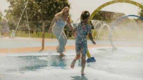 Willen Lake A girl wearing a flowery swimsuit, which covers her legs and arms too, is seen from behind running into water fountains at a splash park. A woman who is blurred out is seen running towards her.