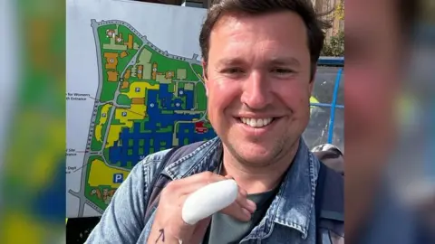 Steve Race is smiling at the camera. He has his finger bandaged and is standing outside a hospital
