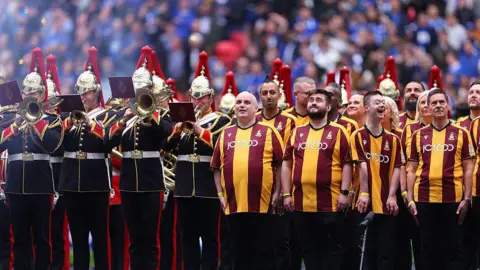 A mixed gender and aged group of people wearing Bradford City FC branded outfits, pose together in a football stadium. They stand alongside a brass band in military uniform.