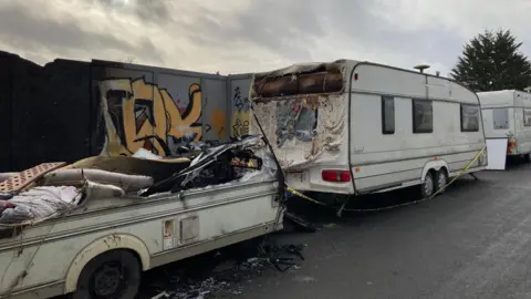 A street in an industrial area of Bristol. One side of the road is lined with caravans, with one completely ravaged by fire. 