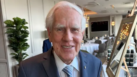 A white haired man smiles to the camera. He wears a dark blue suit, white shirt and blue tie. On his left lapel is a small gold pin. Behind him are rows of tables with white cloth over them, paired with grey chairs. Over his right shoulder is a tall green house plant. Over his left shoulder is a mirror with a cream frame.