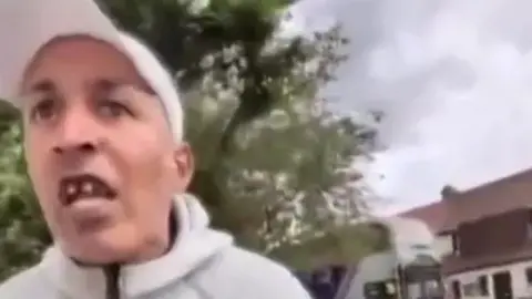Phone footage of a man in a white cap looking angry, leaning towards the person holding it.