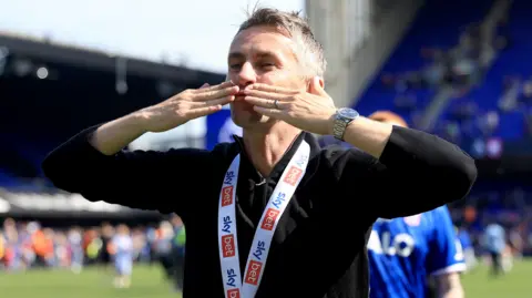 PA Media A man on a football stadium pitch blows kisses to the crowds. He has short grey hair and wears a black jumper with a medal around his neck.