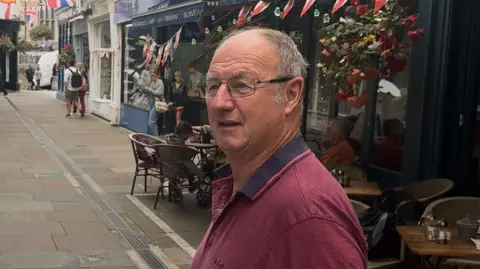 Mick Prigent is pictured. He is standing in front of a café wearing glasses and a burgundy shirt. 