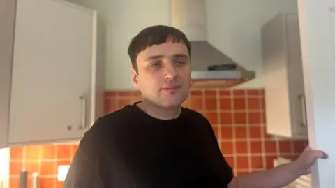 Mid-shot of Timothy Jones standing in a kitchen, his left hand holding onto a white door. He has brown hair and a black t-shirt.